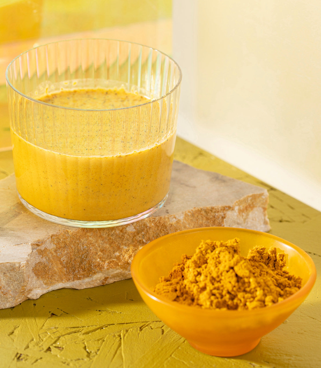 Glass of organic golden milk next to a bowl of turmeric powder on a textured yellow background.