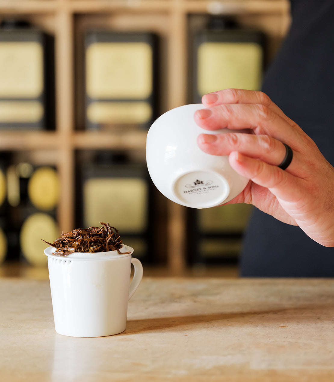 Harney & Sons logo tasting cup set with loose tea being poured into a white cup on a wooden surface.