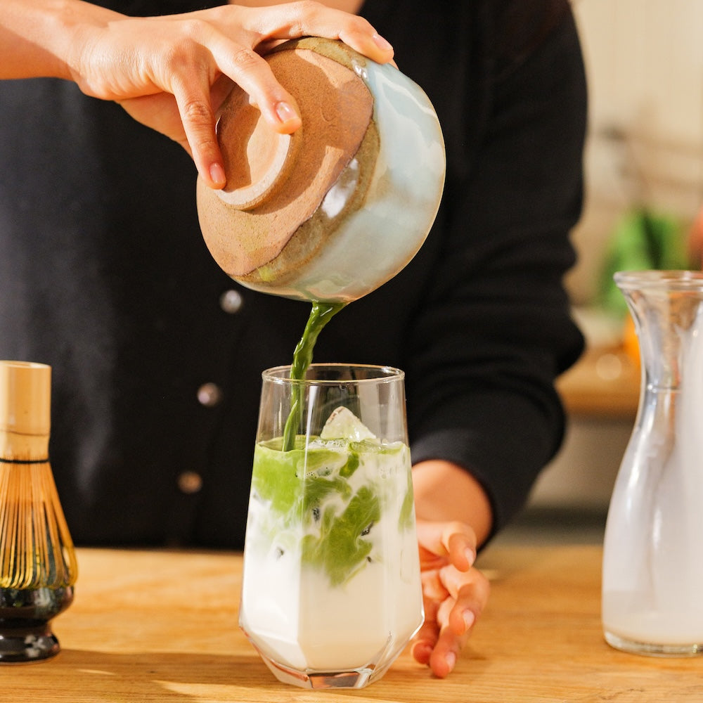 A ceramic bowl of matcha being poured into a glass full of milk and ice, creating a latte, set on a kitchen counter.