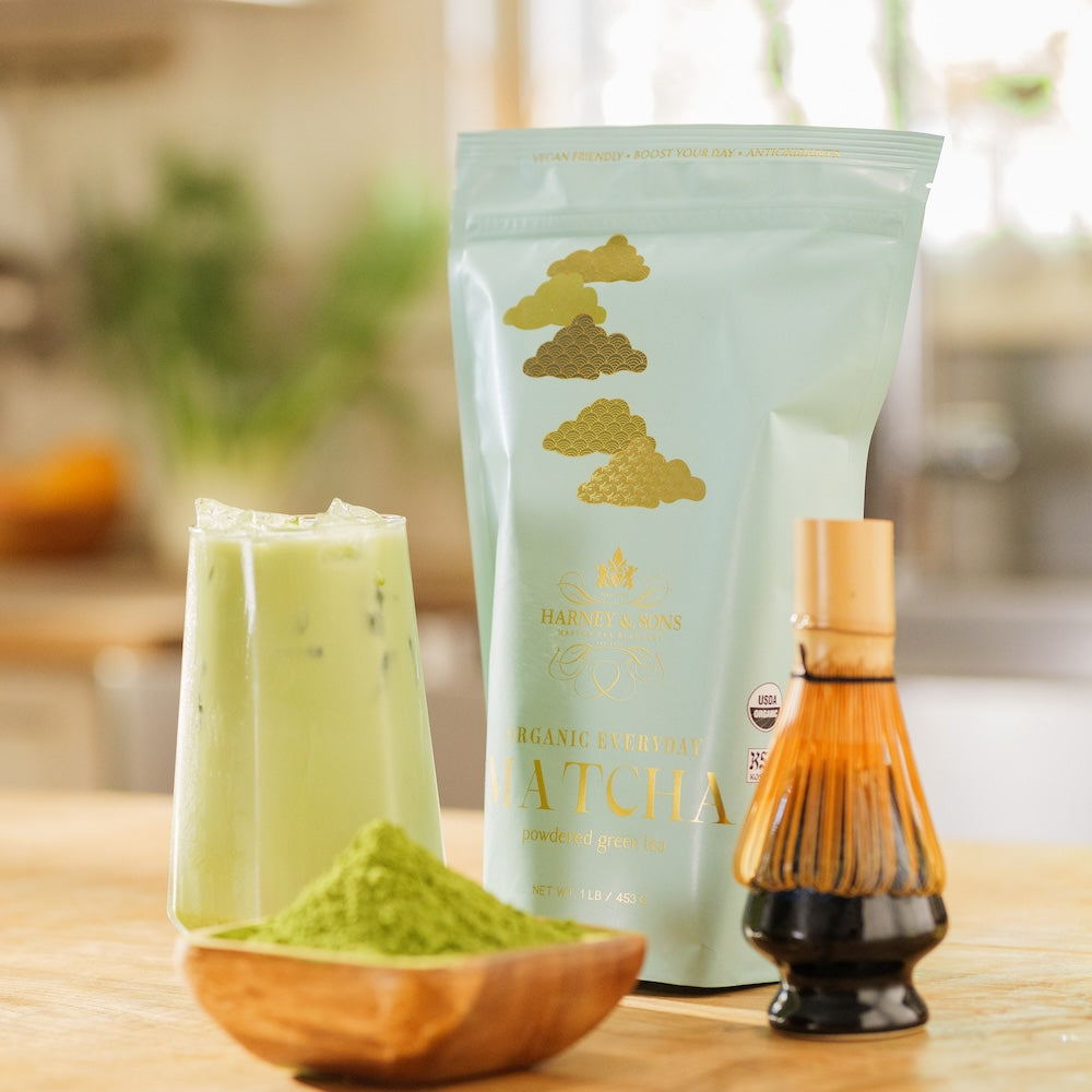 A bag of Organic Everyday Matcha on a counter with a latte, green powder in a wooden bowl, and a whisk on a ceramic stand.