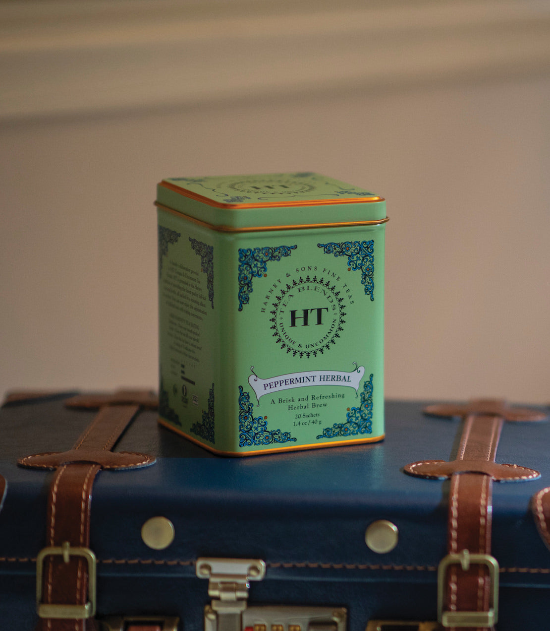 Green tin of Peppermint Herbal tea sachets, featuring decorative design, placed on a vintage suitcase.