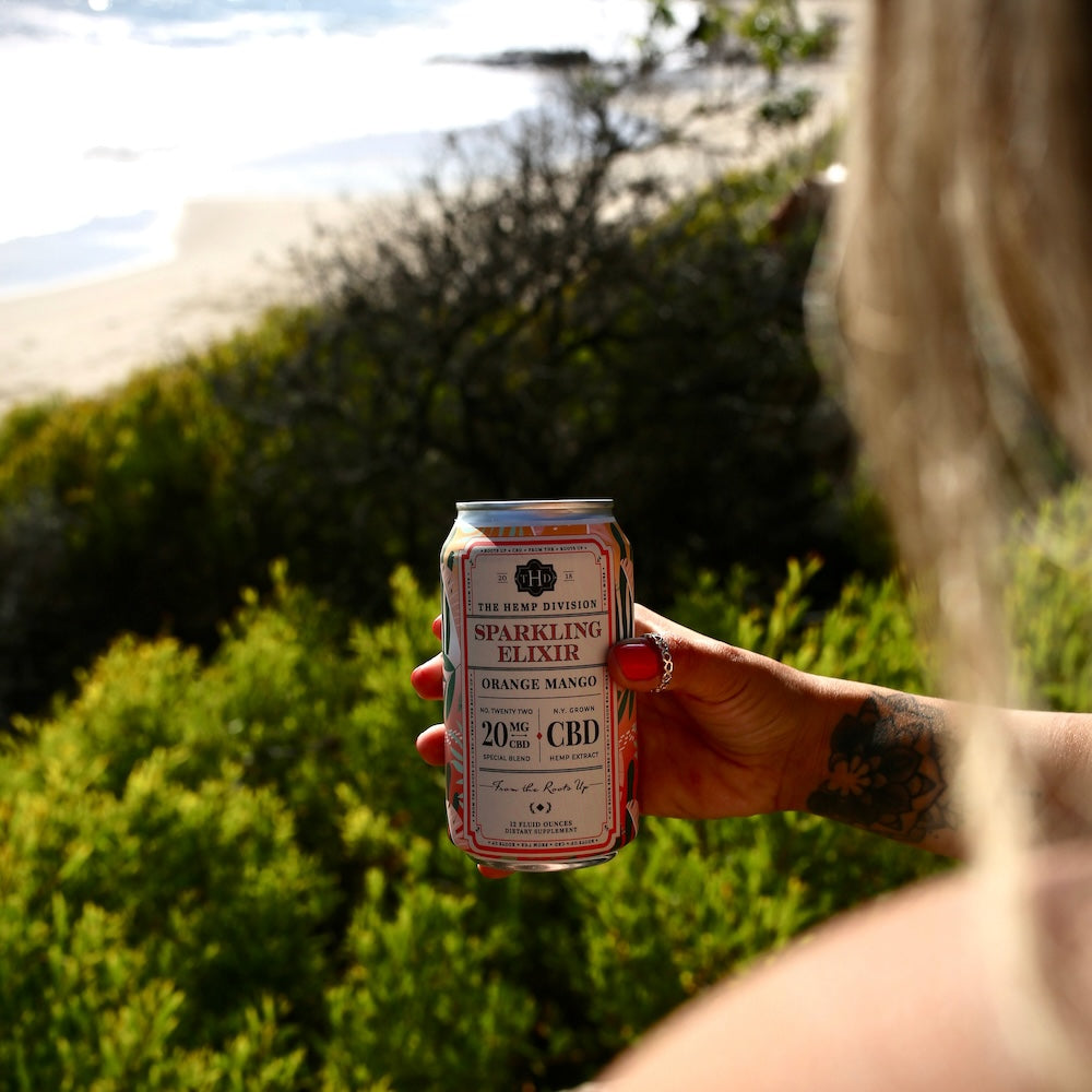 A hand holding a can of Sparkling Elixir Orange Mango with 20 MG CBD, set against a beach backdrop.