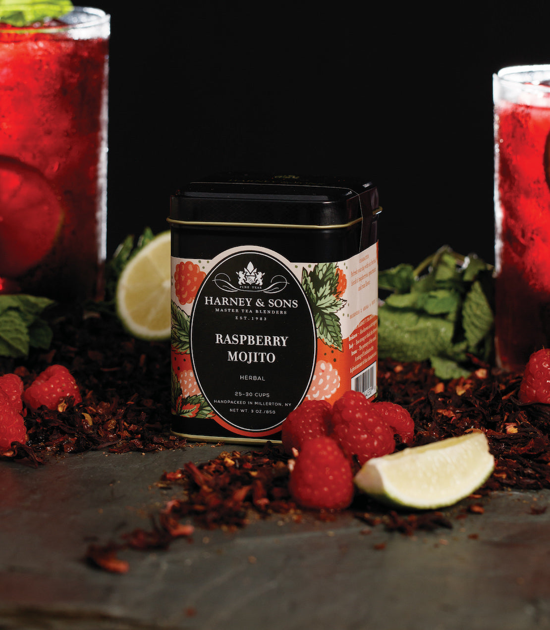 Raspberry Mojito tea by Harney & Sons, surrounded by fresh raspberries, mint, and lime, with drinks in the background.