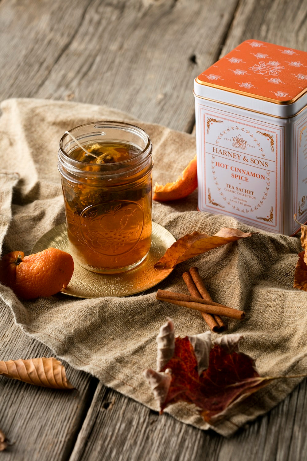 A glass jar of Harley & Sons Hot Cinnamon Spice tea sits on a small gold dish beside an orange-patterned tin box, arranged on a rustic linen cloth with cinnamon sticks, dried leaves, and orange slices on a wooden table.