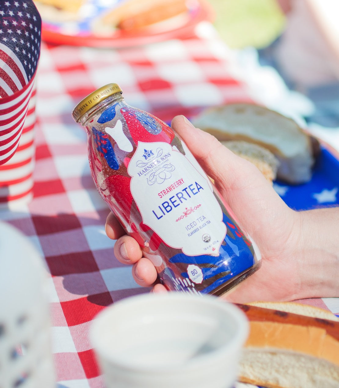 Hand holding a bottle of Strawberry Libertea iced tea on a picnic table with an American flag in the background.