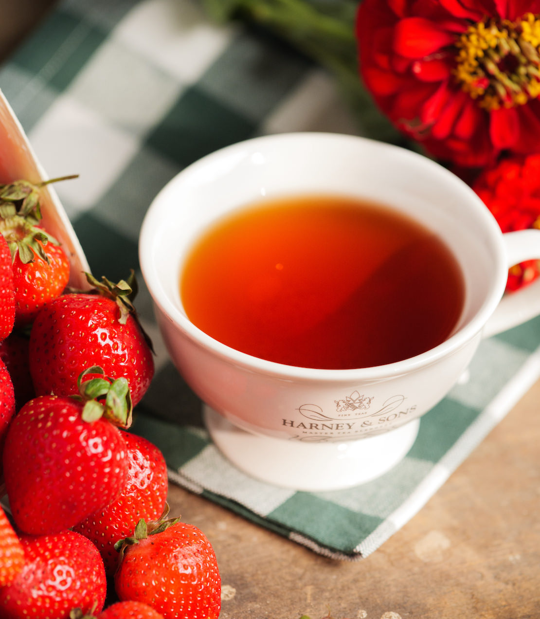 White cup of tea with a Harney & Sons logo, surrounded by strawberries on a wooden surface.