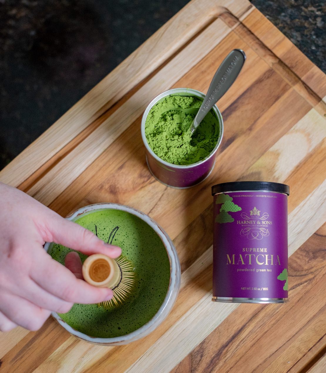 Matcha green tea preparation on a wooden surface with a tin of supreme matcha and a whisk.