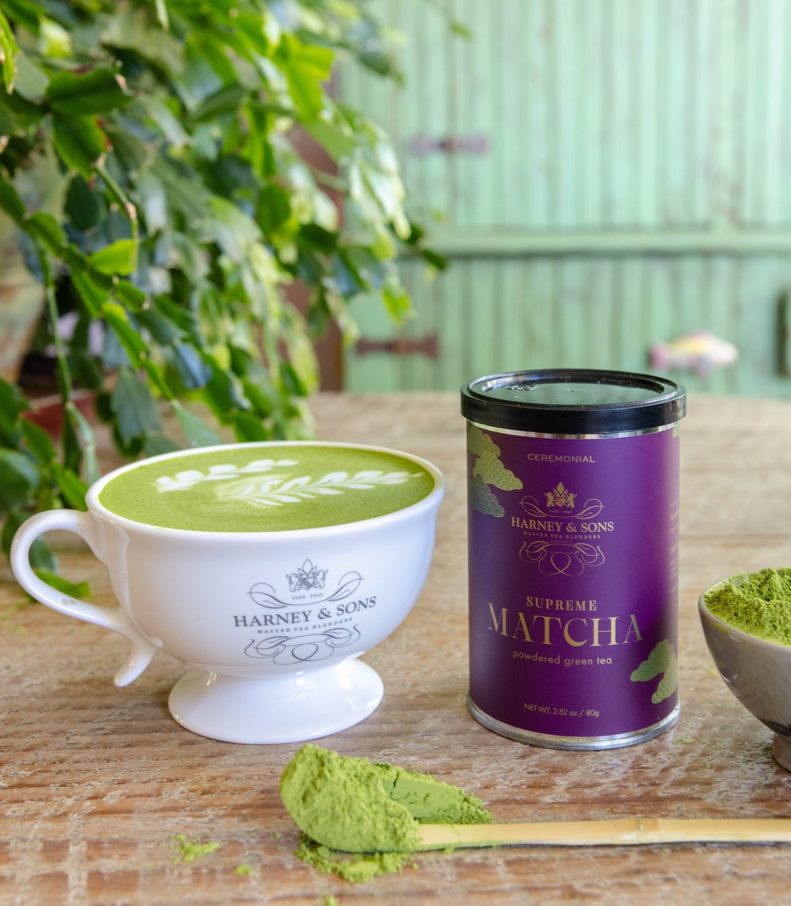 A Supreme Matcha latte with leaf latte art beside a tin of Supreme Matcha and a bamboo scoop of matcha powder on a wooden table.