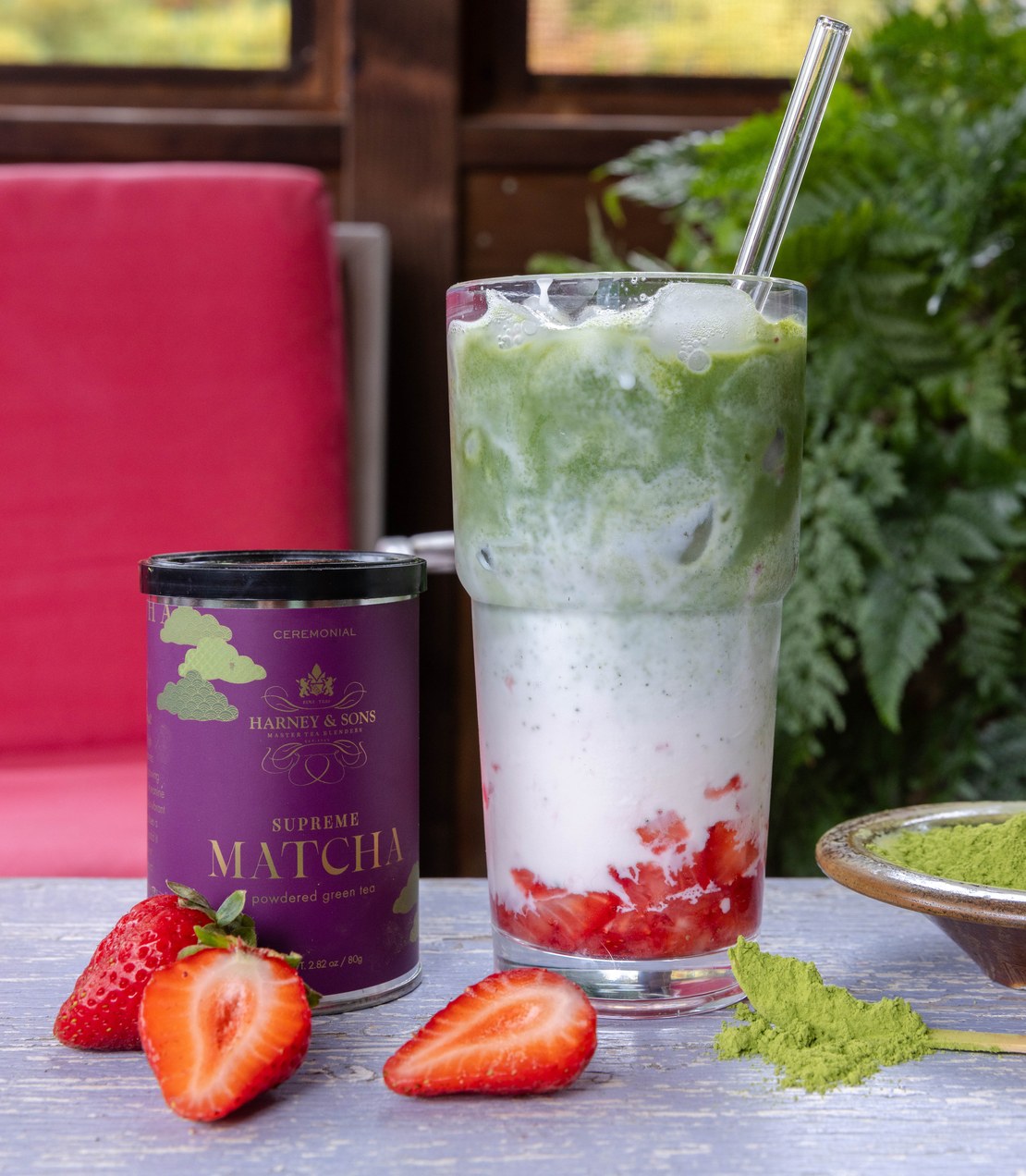 A strawberry iced Supreme Matcha latte with a glass straw beside a tin of Supreme Matcha, fresh strawberries, and a bamboo scoop of matcha powder.