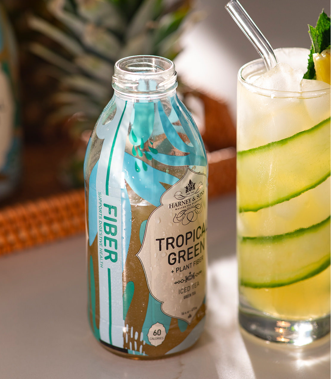 Bottle of Tropical Green + Plant Fiber Iced Tea next to a glass garnished with cucumber and mint.