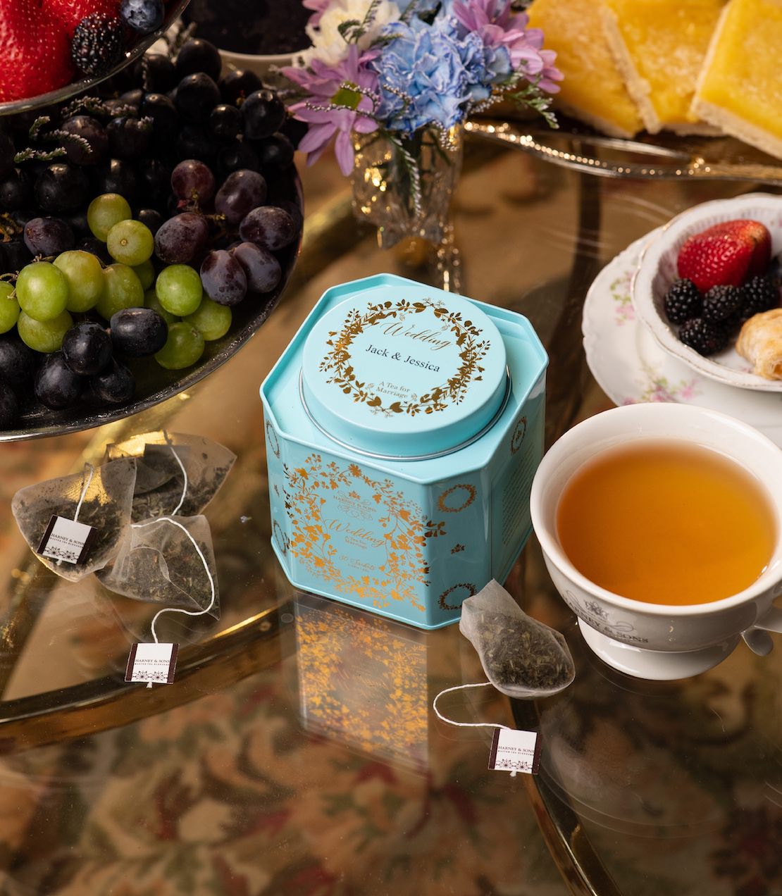 Personalized wedding tea tin featuring gold floral design, containing 30 sachets on a table surround by treats and flowers.