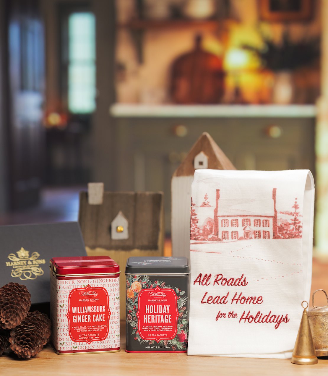 A holiday gift set on a wooden surface featuring a red tea towel labeled 'all roads lead home for the holidays', two decorative metal tins, a dark gift box, pinecones, and a blurred warm-toned indoor background.