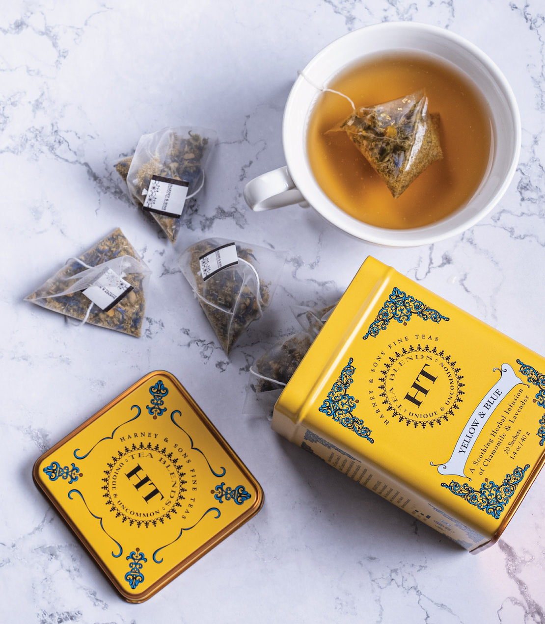 Yellow and blue tin of HT tea sachets with a cup of brewed tea and sachets on a marble surface.