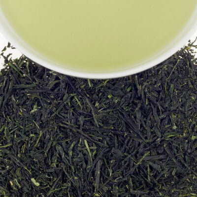Close-up of dark, dried loose tea leaves with a light green herbal tea in a white cup above.
