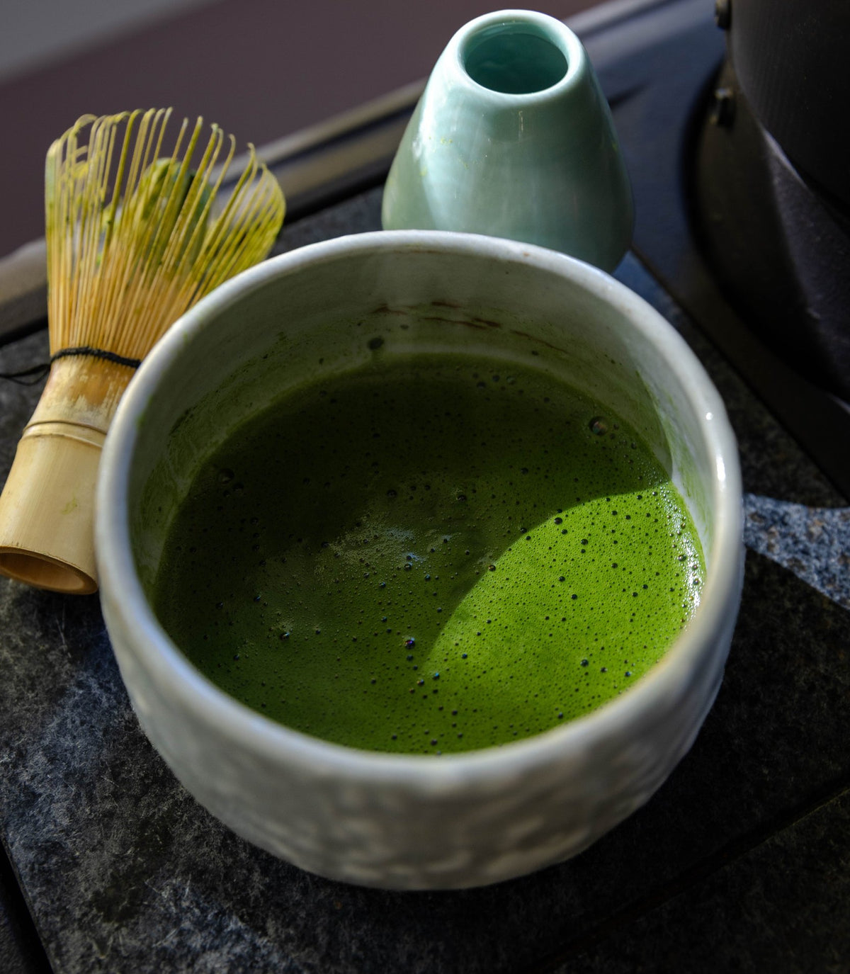 A bowl of whisked matcha beside a bamboo whisk, seen from above on dark stone.