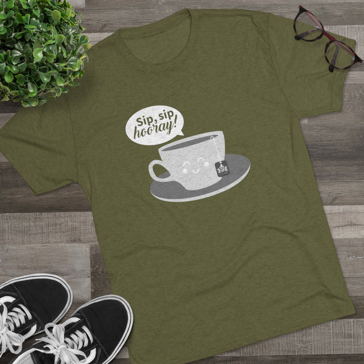 Olive green graphic tee featuring a coffee cup design with the text "Sip, sip, hooray!" on a wooden background.
