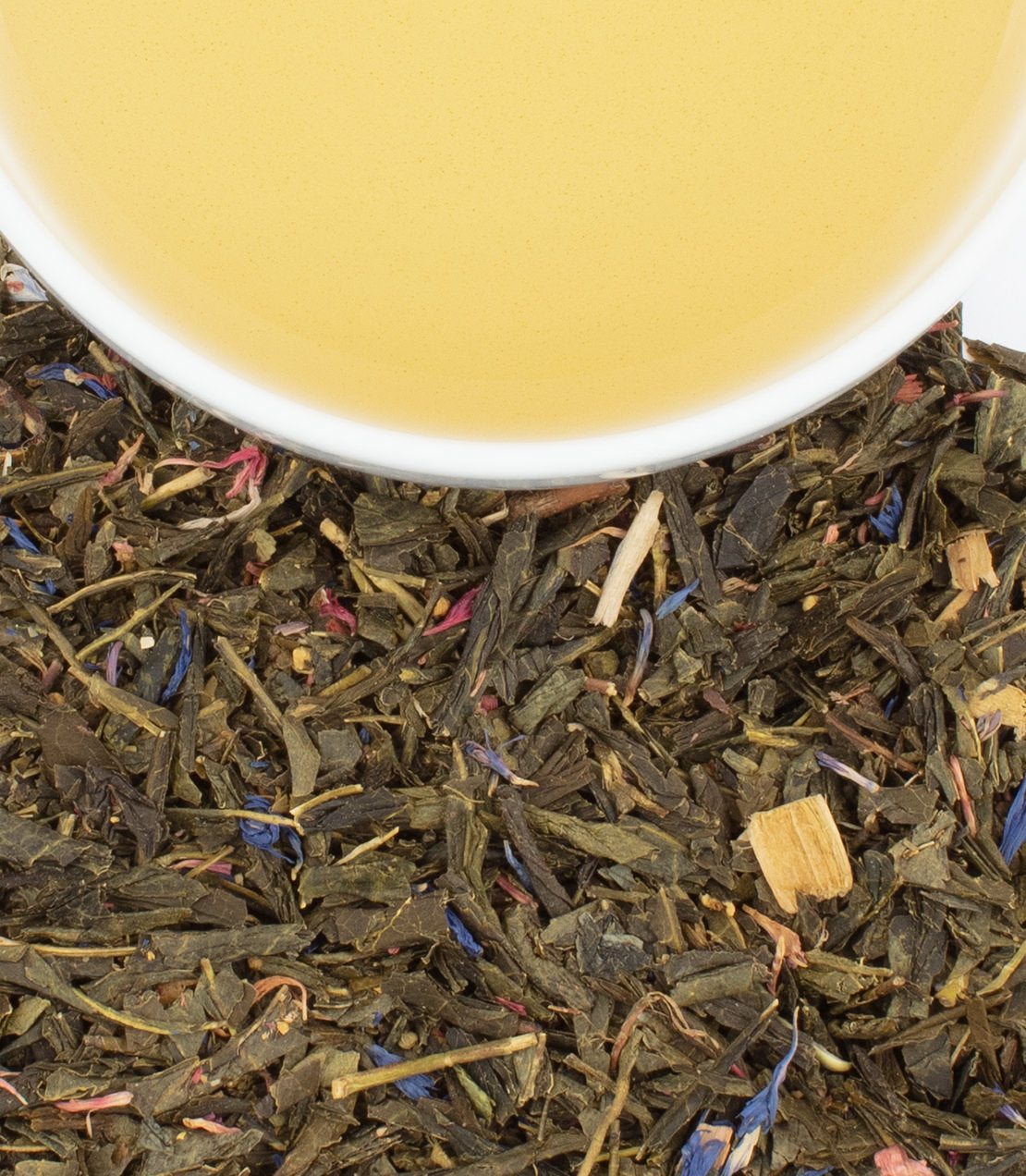 A close-up of loose green tea leaves with colorful flower petals, next to a cup of brewed tea.