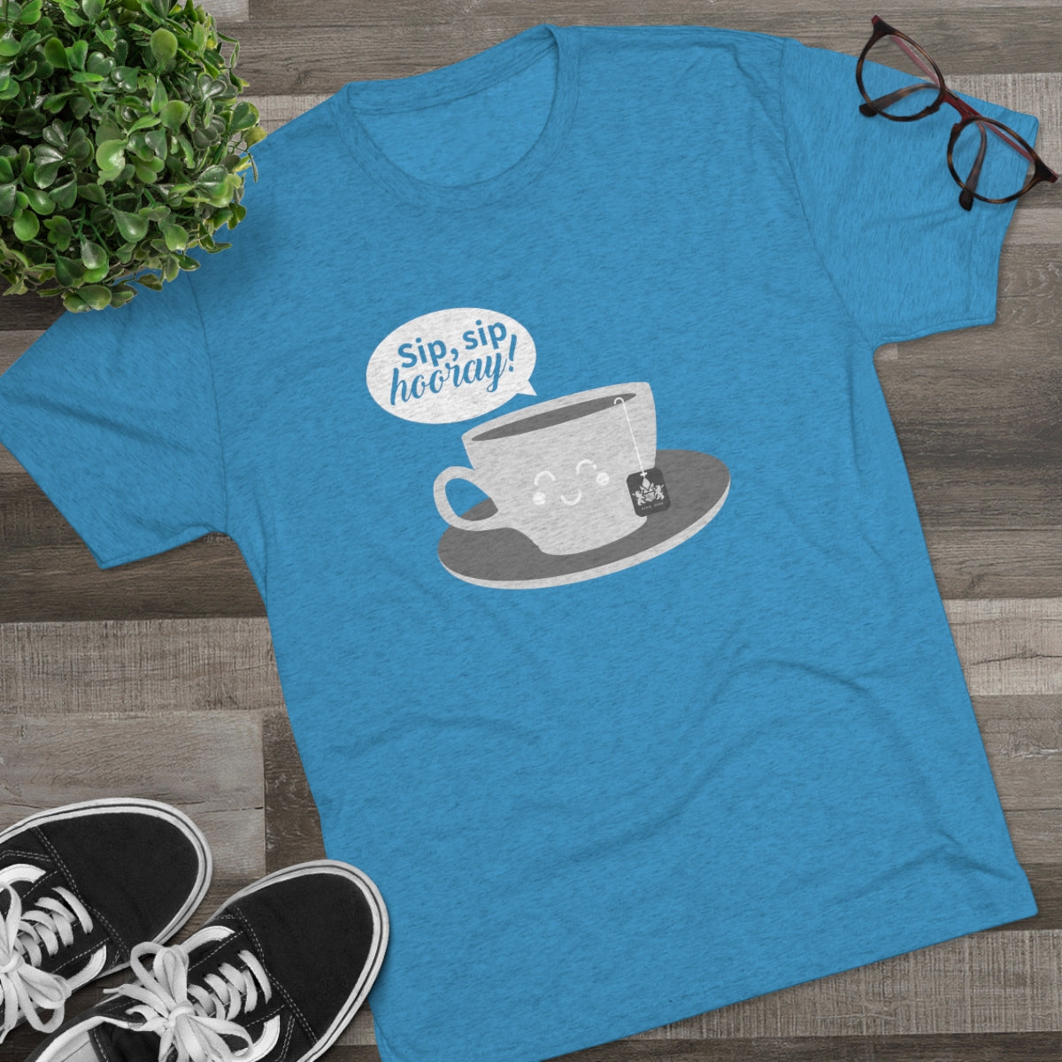 Blue graphic tee featuring a smiling teacup and the text "Sip, sip, hooray!" on a wooden surface with glasses and sneakers.