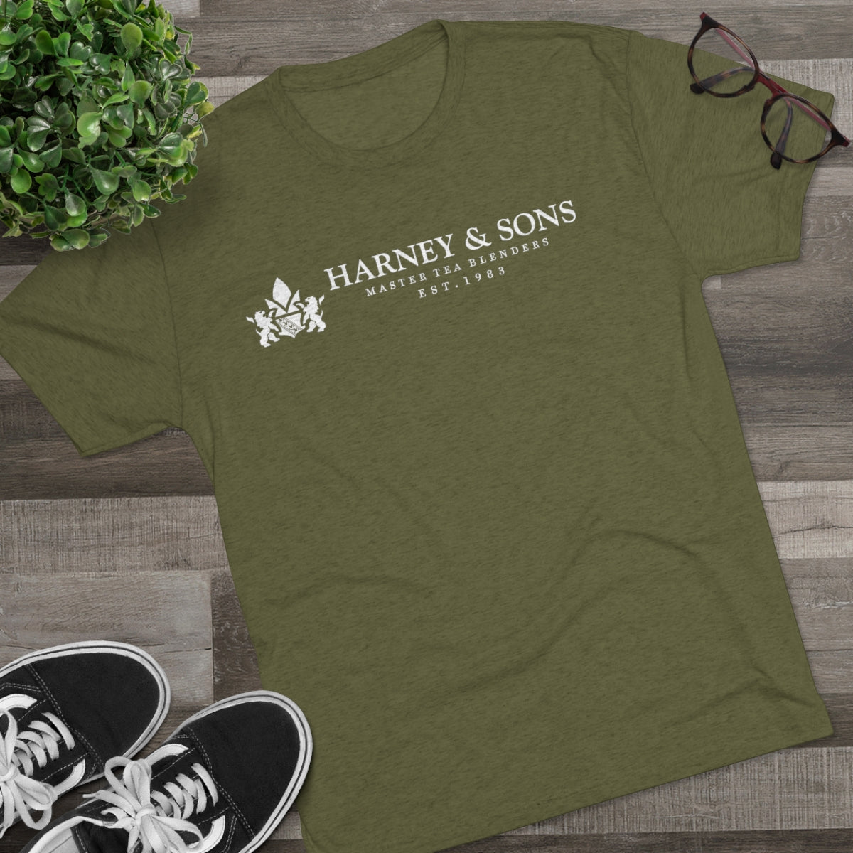 Olive green graphic tee featuring "Harney & Sons" logo, established 1983, with black sneakers and glasses nearby.