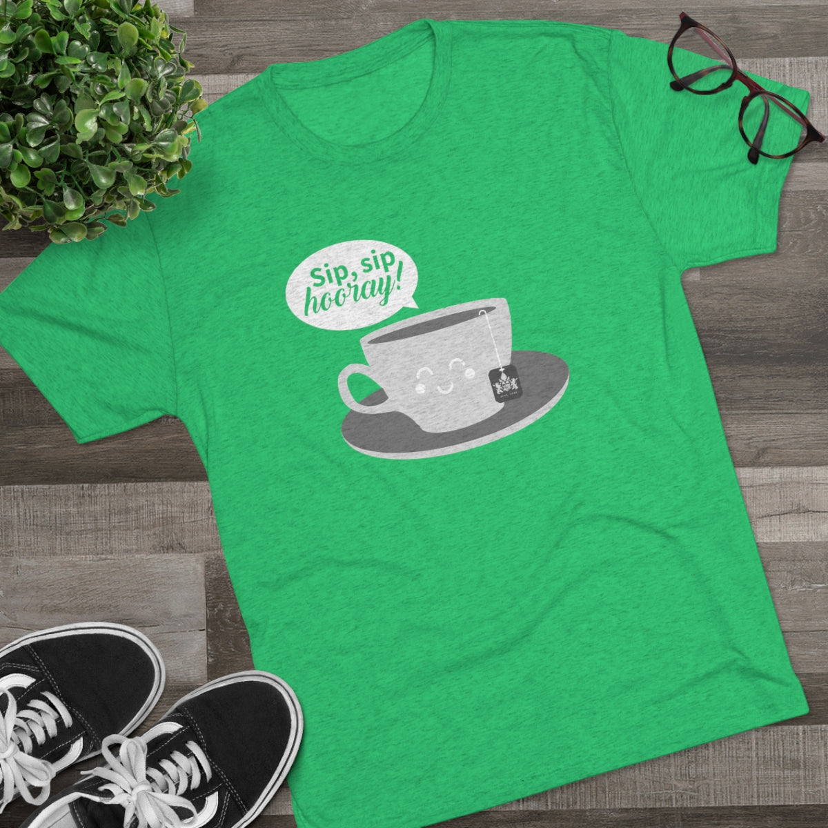 Green graphic tee featuring a smiling teacup and the text "Sip, sip, hooray!" on a wooden background.