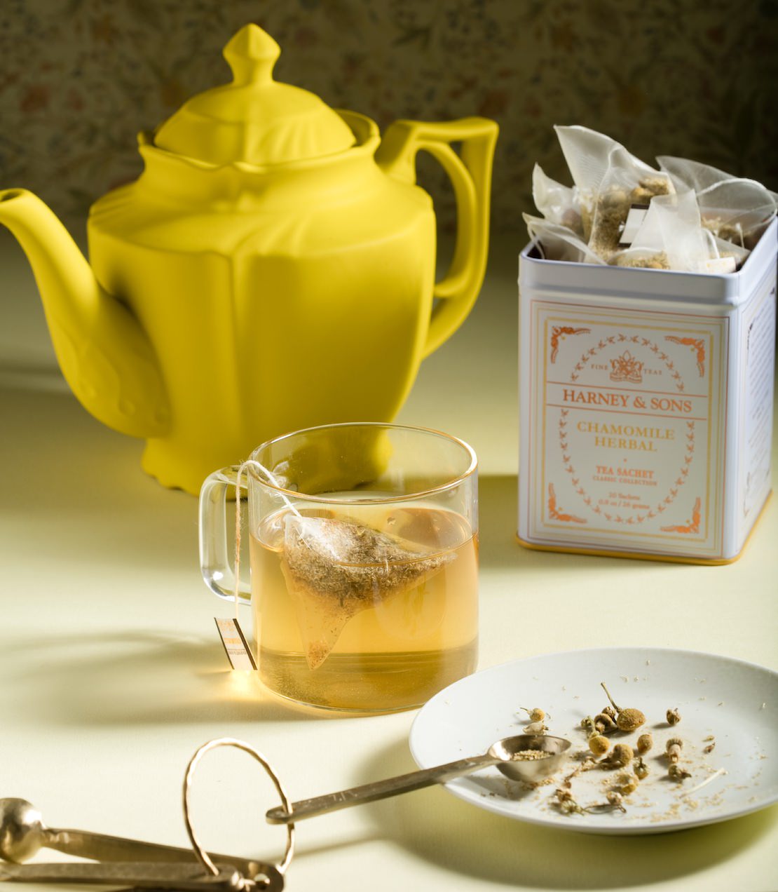 Chamomile Herbal tea sachets in a classic tin, with a brewed cup and loose chamomile flowers on a plate.