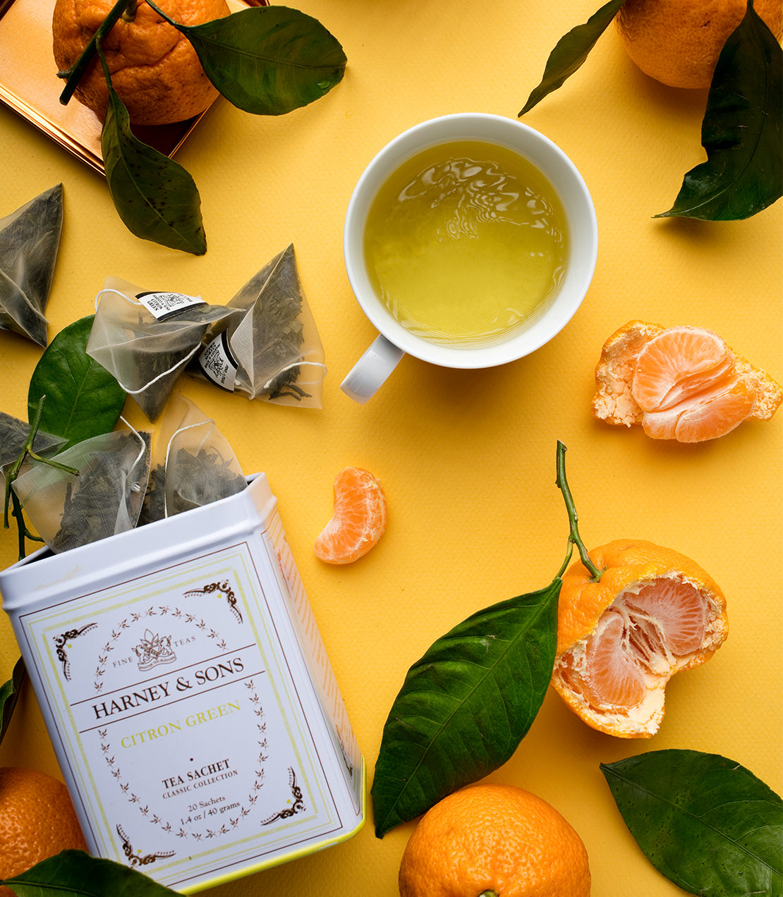 Citron Green tea sachets in a classic tin, surrounded by fresh citrus fruits and a cup of brewed tea.
