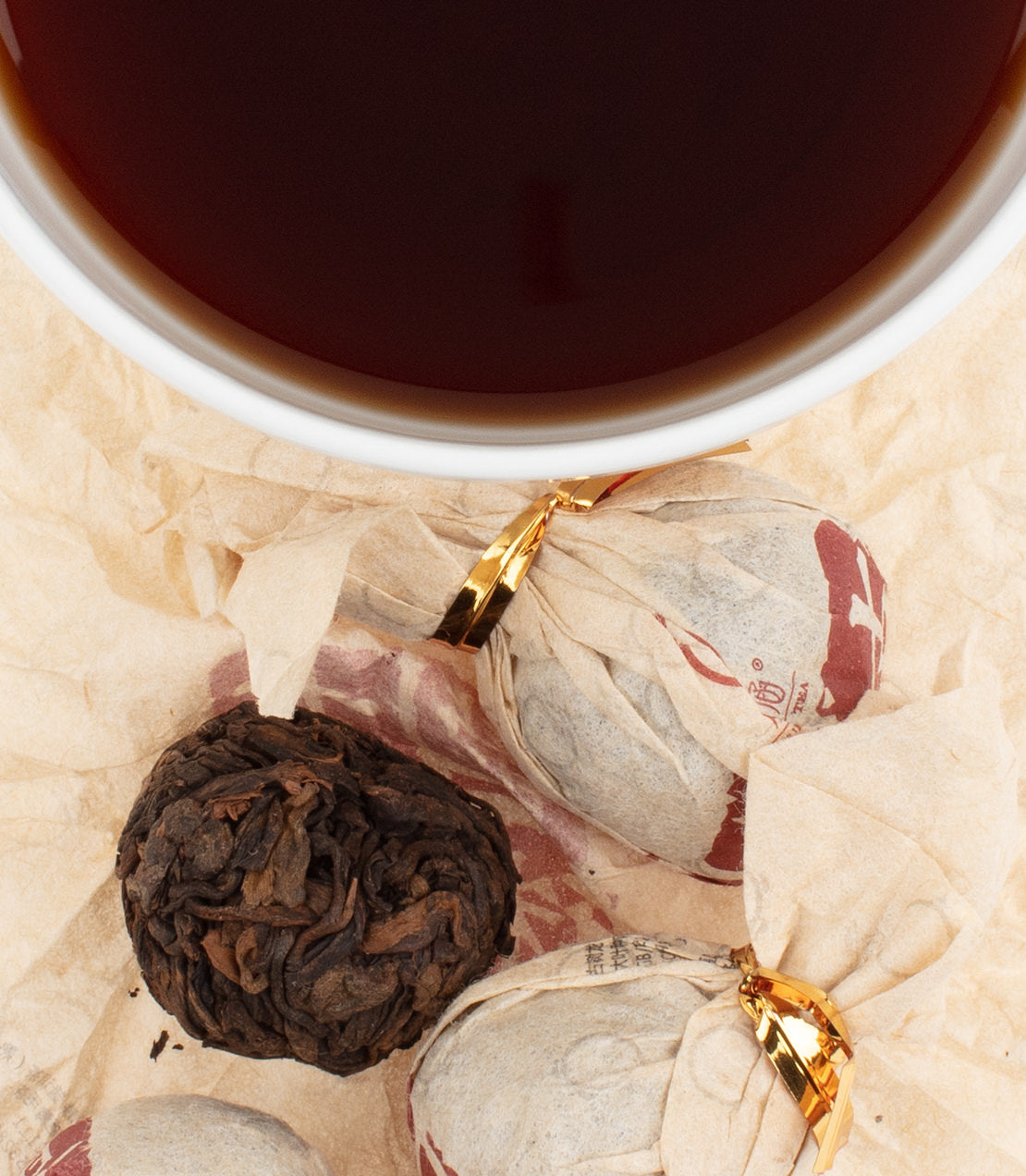 Ripe Pu-Erh tea balls beside a cup of dark brewed tea on crumpled paper. Rich flavor and aroma await.