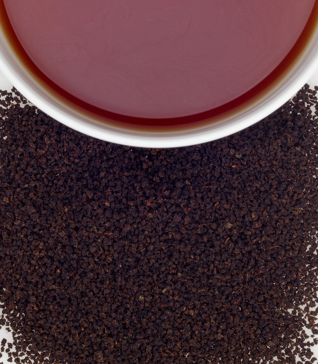 CTC Assam tea in a white cup, surrounded by loose black tea granules on a white background.