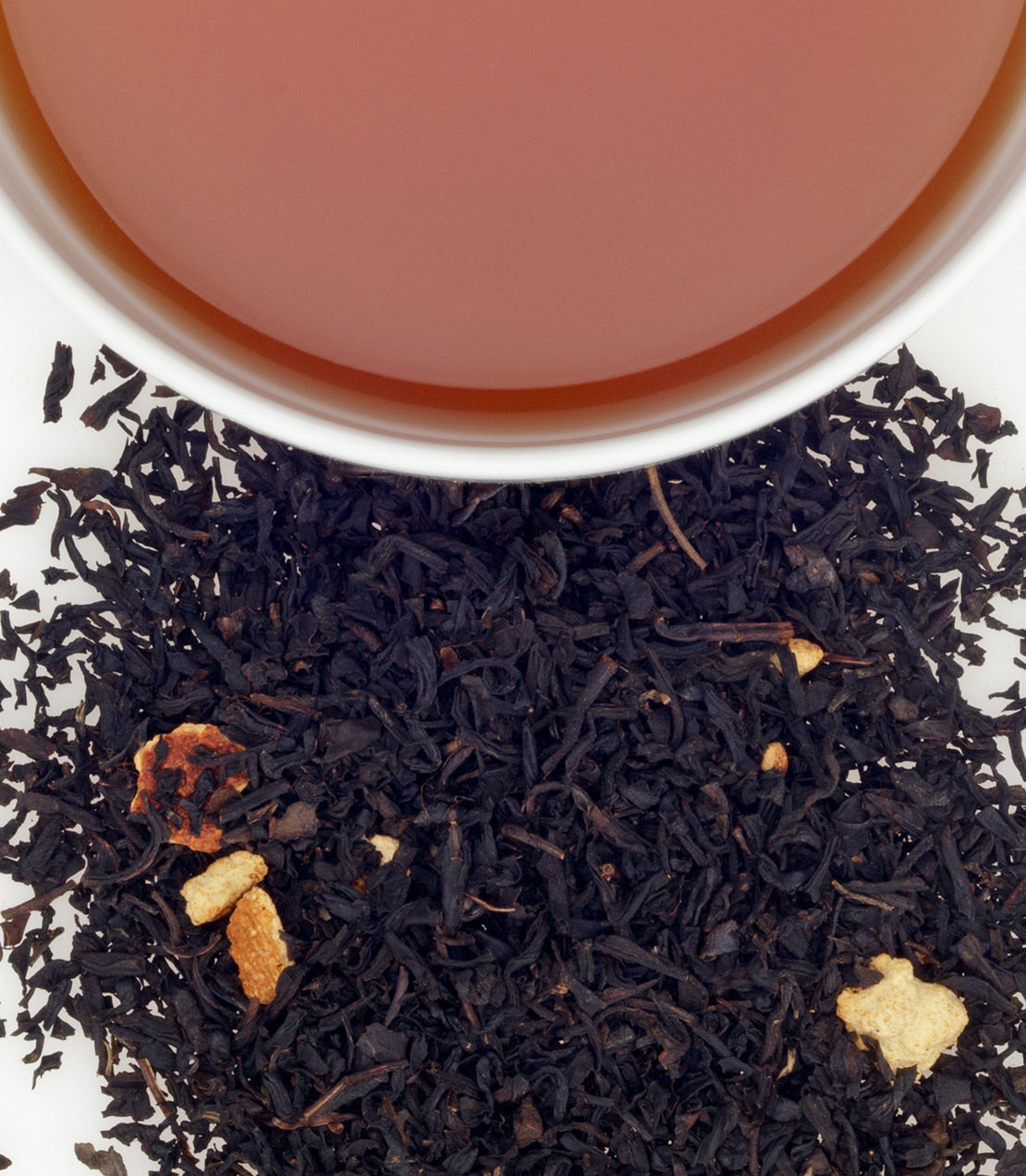 A close-up of loose black tea leaves with dried orange pieces beside a cup of brewed tea.