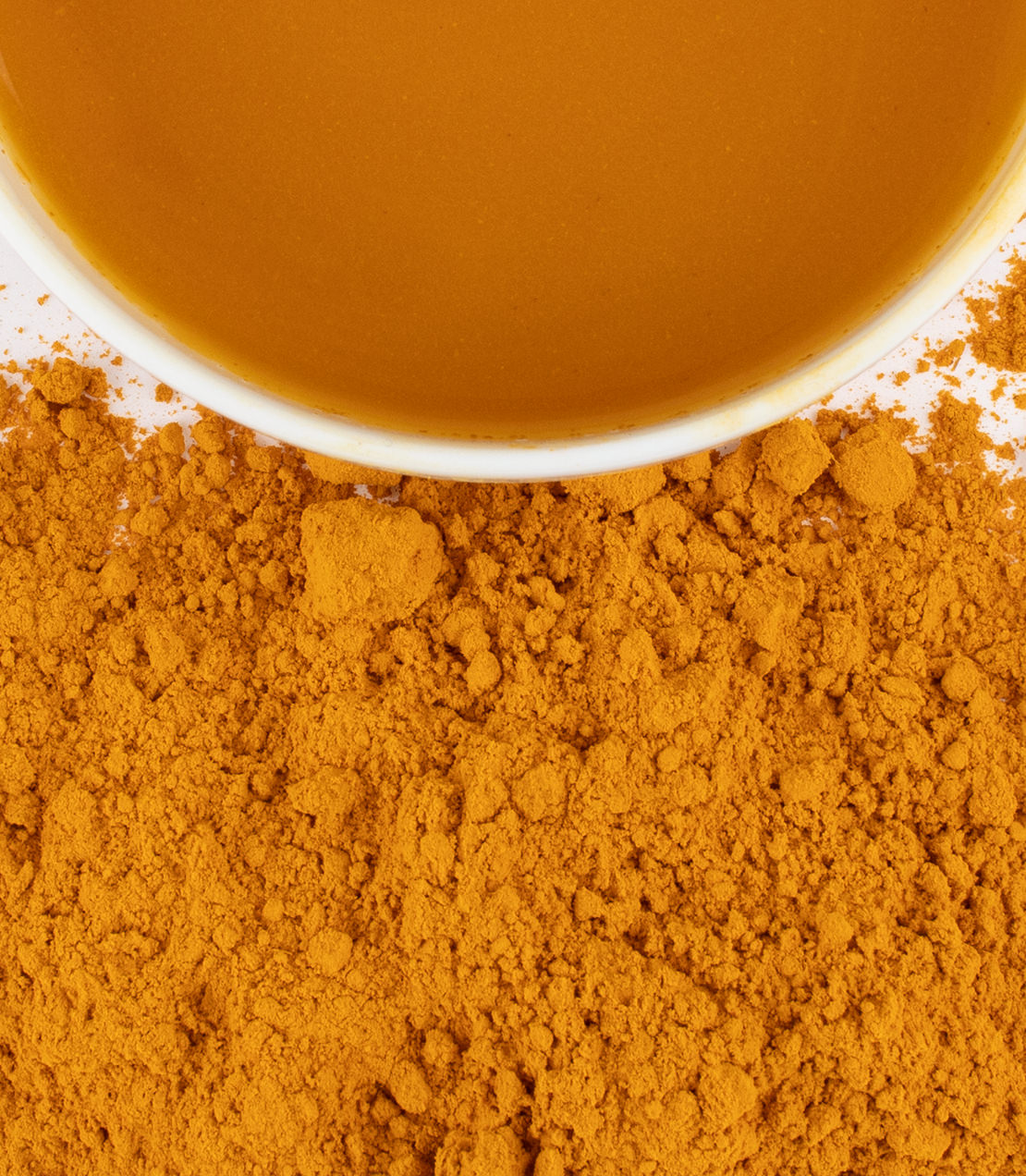 Organic turmeric powder scattered around a white bowl filled with turmeric paste, showcasing its vibrant yellow-orange color.