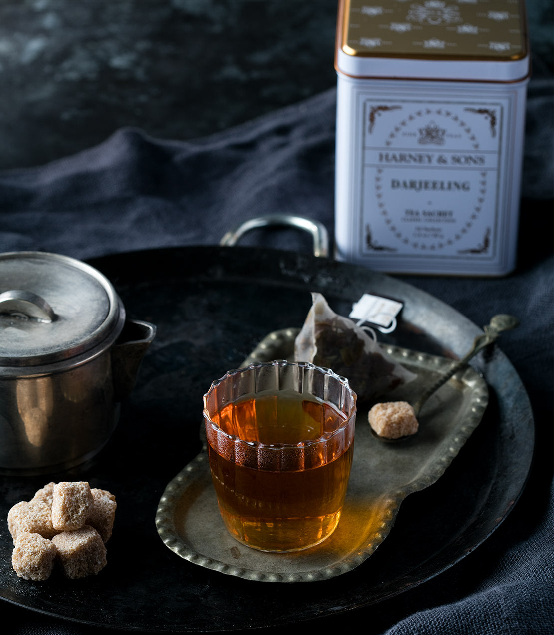 Classic Darjeeling tea sachets in a decorative tin, served with a glass of brewed tea and sugar cubes on a tray.