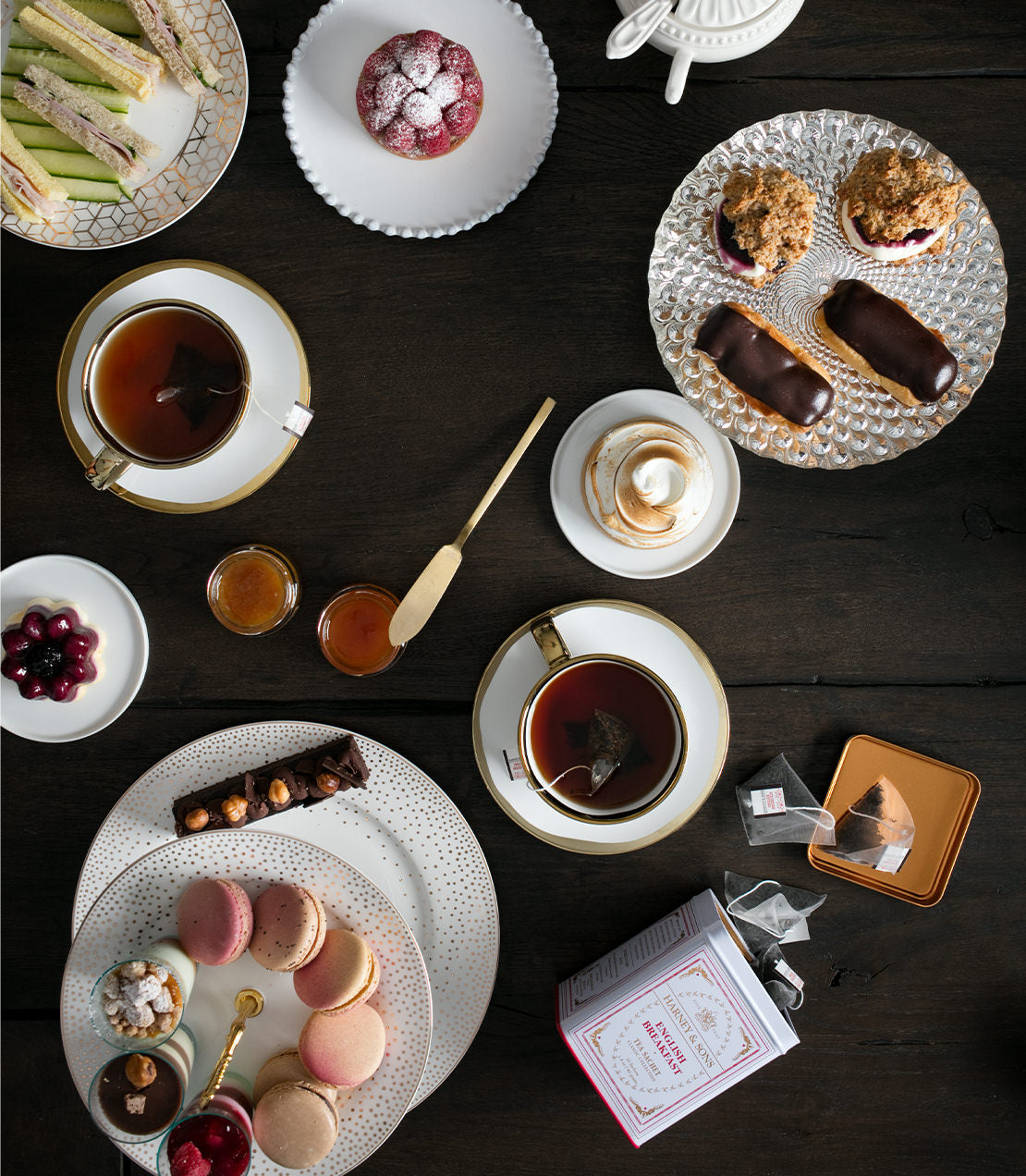 A classic tin of English Breakfast tea sachets surrounded by elegant tea cups, pastries, and desserts on a wooden table.