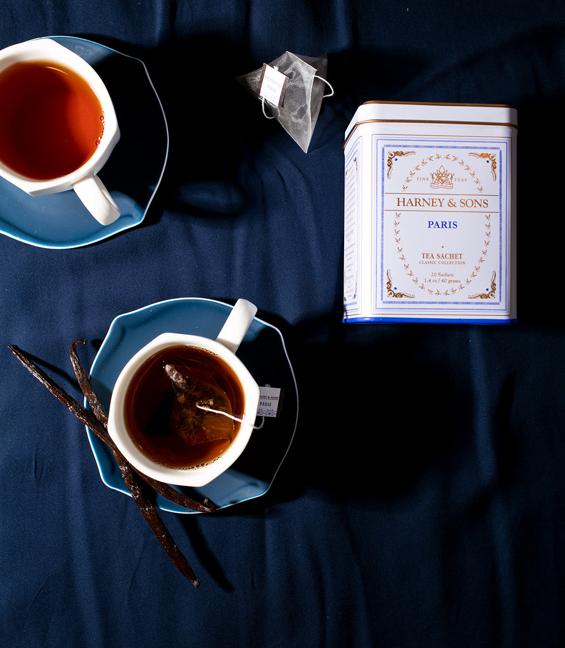 Classic tin of 20 Paris tea sachets by Harney & Sons, with brewed tea in cups and vanilla pods on a dark background.