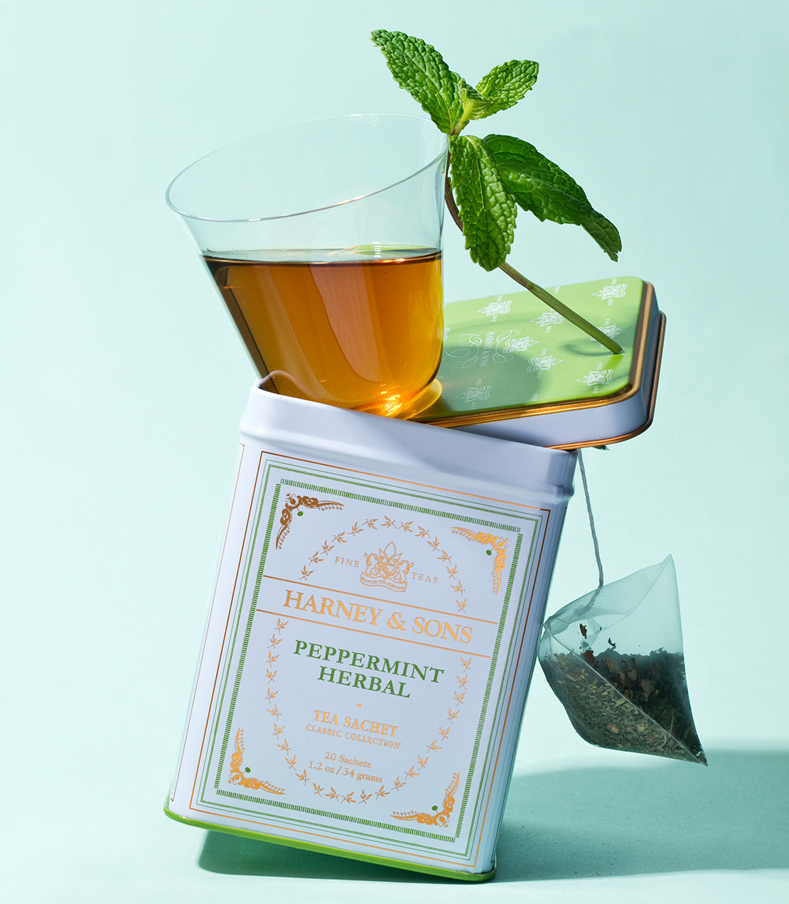 Classic tin of Harney & Sons Peppermint Herbal tea sachets with a glass of brewed tea and fresh mint leaves.