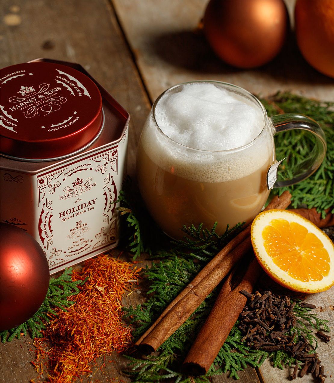 Warm cup of holiday tea with frothy milk, surrounded by spices, orange slice, and festive decorations.