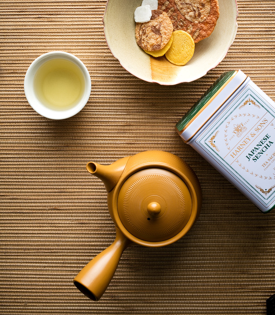 Classic tin of Japanese Sencha tea sachets beside a teapot and a bowl of snacks on a textured mat.