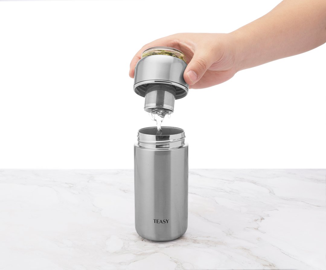 Hand holding the lid of a Teasy insulated flask, pouring liquid into the stainless steel body on a marble surface.