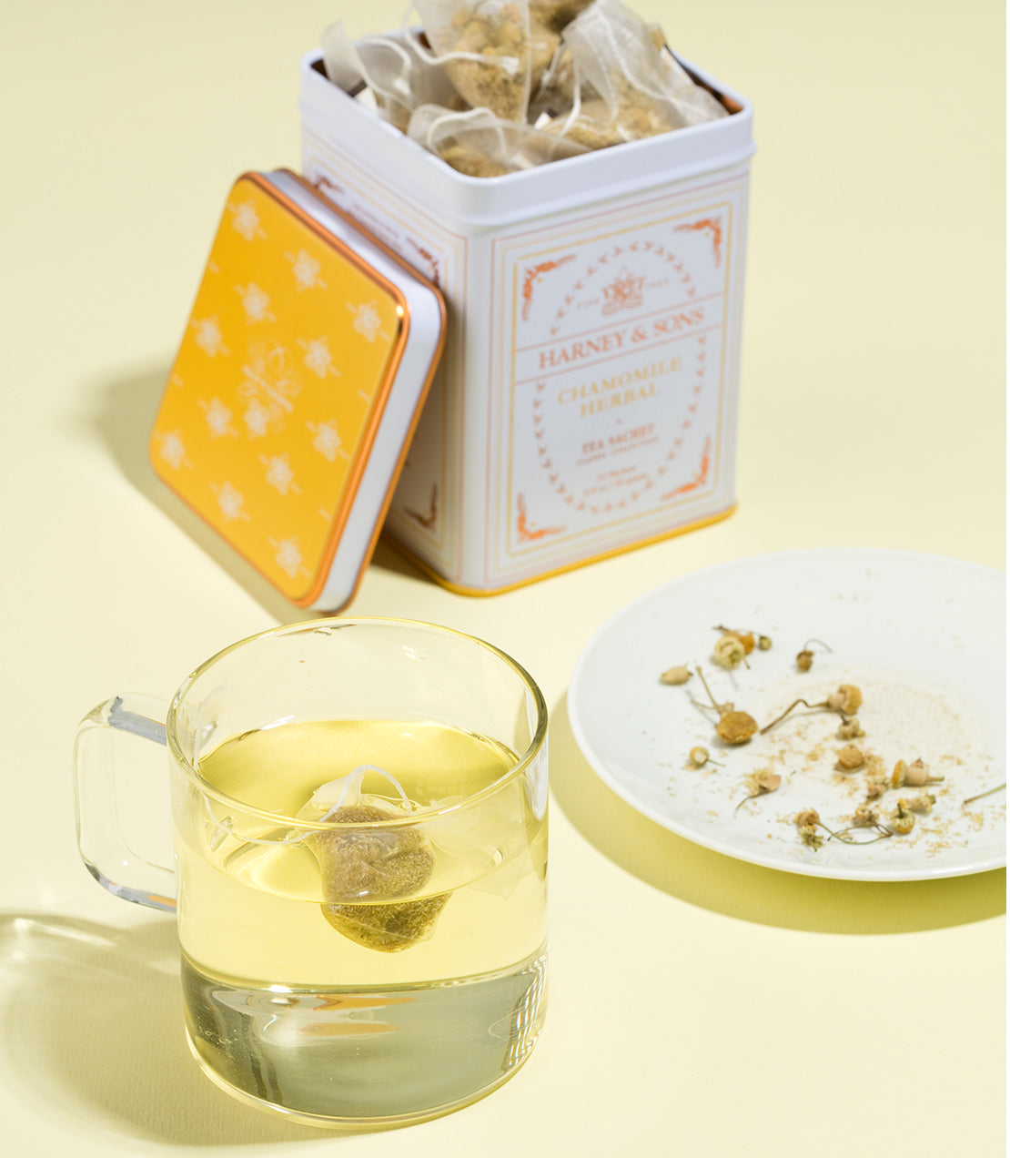 Chamomile Herbal tea sachets in a classic tin, with a brewed cup and loose chamomile flowers on a plate.