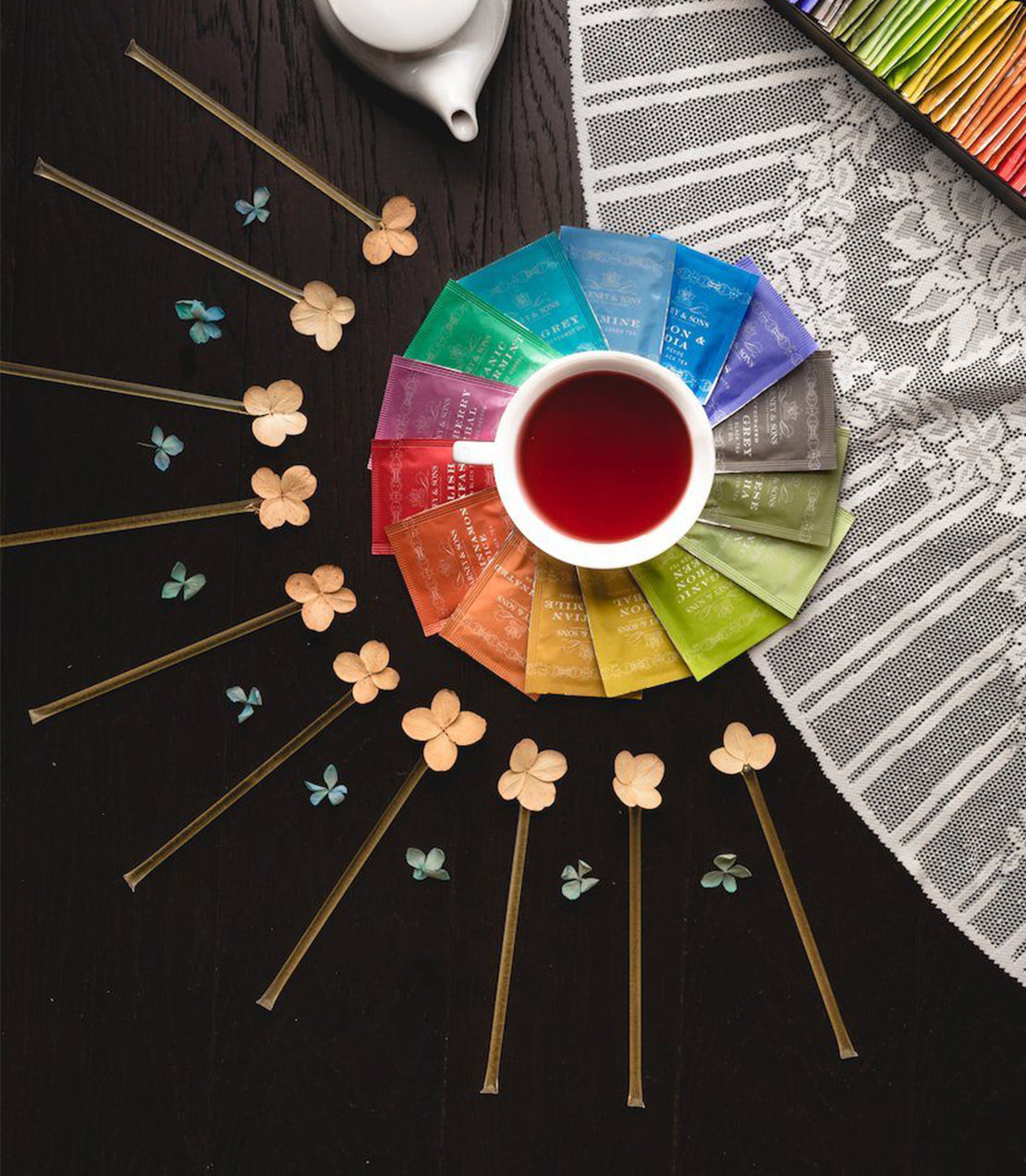 Colorful teabag and honey sampler arranged around a cup of tea, with decorative wooden stirrers and flowers.