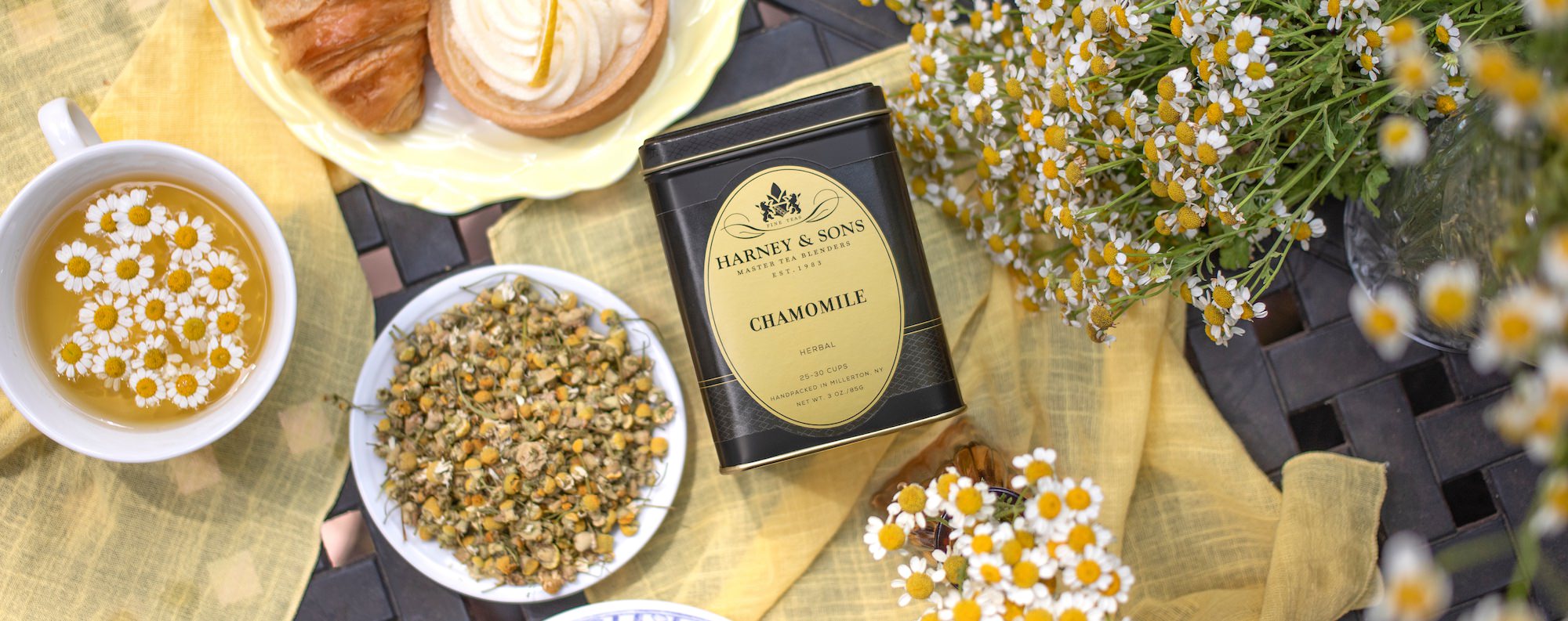 Chamomile tea tin with a cup of tea, pastries, and flowers on a wooden surface.