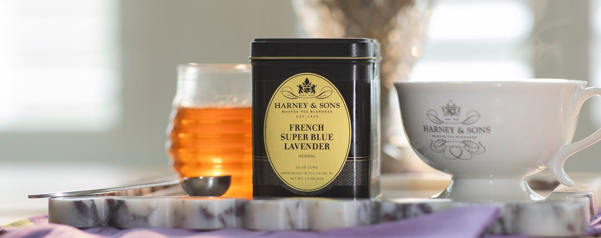 Harney & Sons French Super Blue Lavender tea tin with a cup, tea spoon,  and jar of honey on a marble surface.