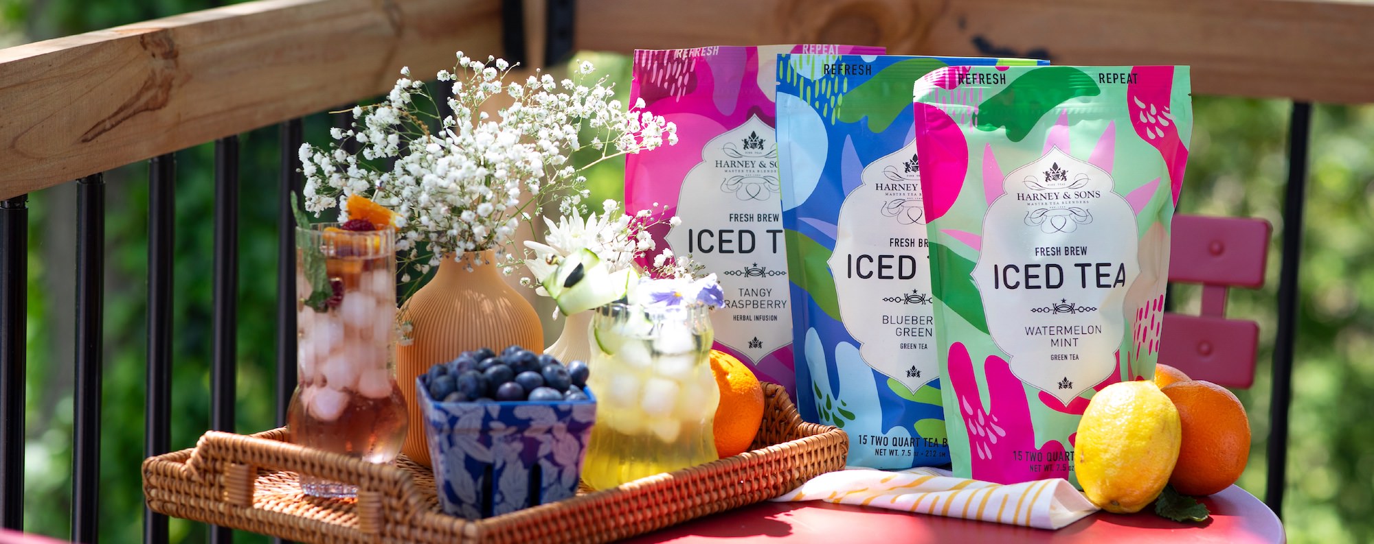 Colorful iced tea packages on a tray with flowers and fruits outdoors. Tangy Raspberry, Blueberry Green, and Watermelon mint are featured. 