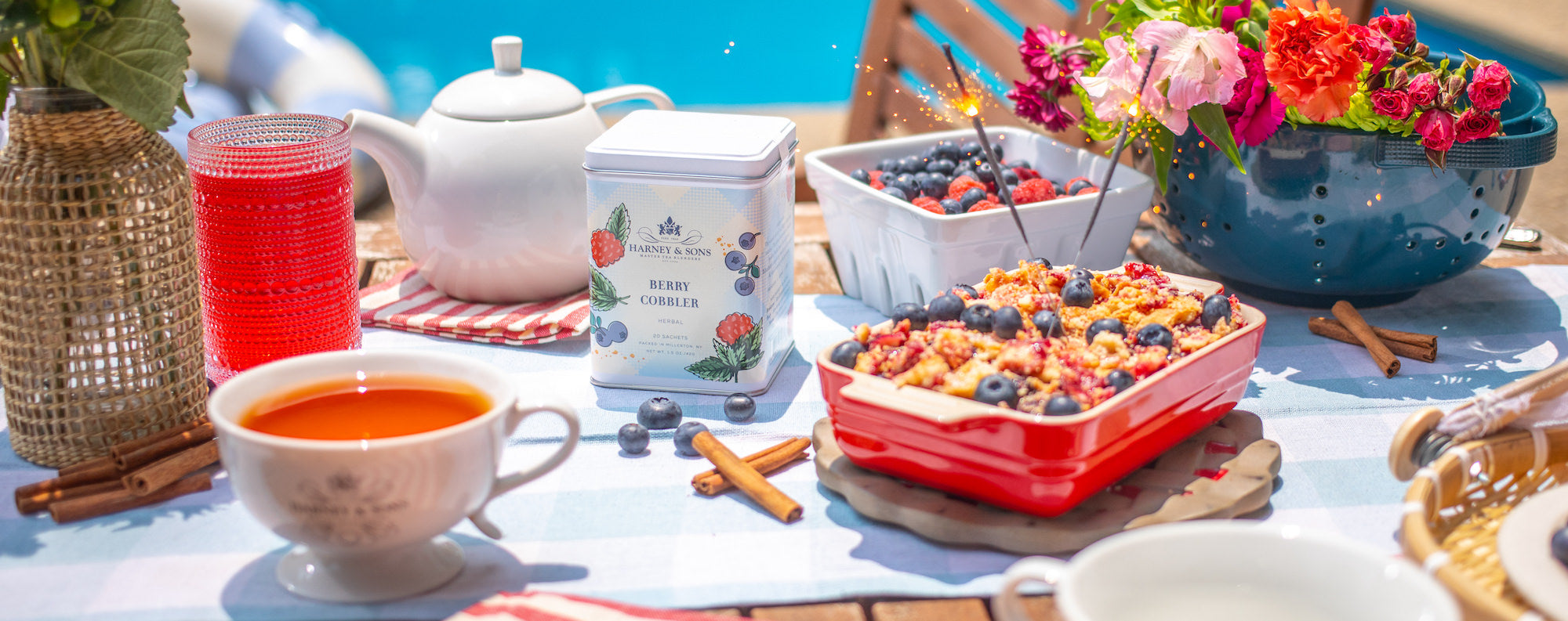 Berry Cobbler sachet tin sitting on a table surrounded by cobbler pie, cups of tea, and fresh fruit. Everything on the table is red, white, and blue colors, suggesting an independence day theme. 
