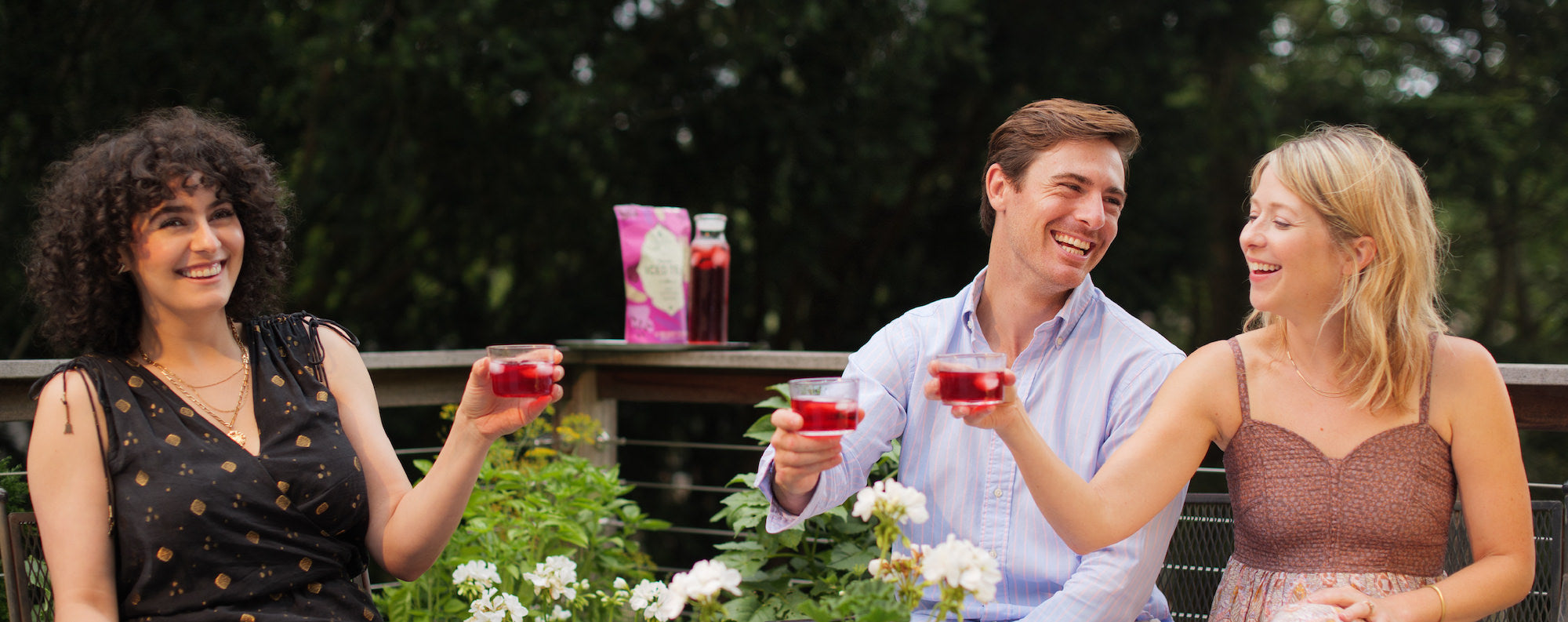 2 women and a man smiling and enjoying Tangy Raspberry Fresh Brew iced tea. They're sitting outside on a deck surrounded by plants and trees. 