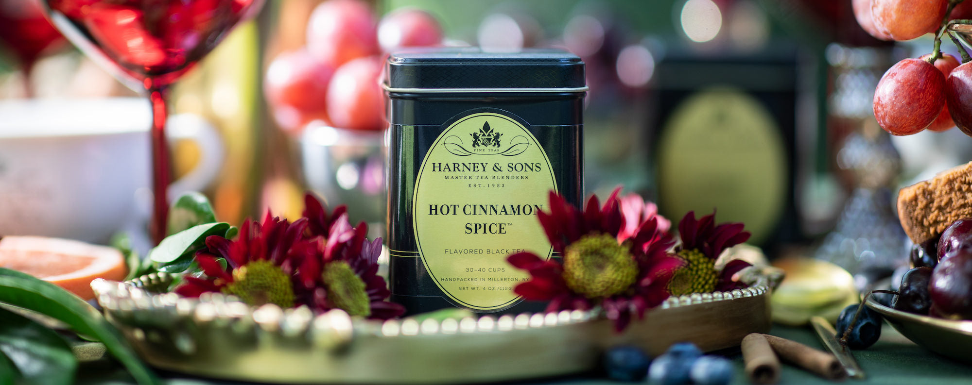 Harney & Sons Hot Cinnamon Spice tea tin on a decorative table with flowers and fruits.