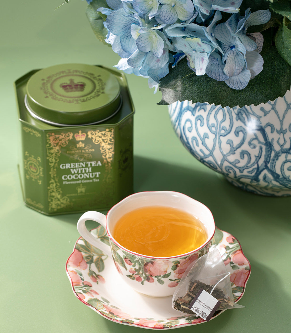 Harney & Sons Fine Teas- Historic Royal Palaces- Green Tea with Coconut- Green tin of 30 sachets- Lifestyle shot with cup of tea and sachet- vase of blue flowers- green background