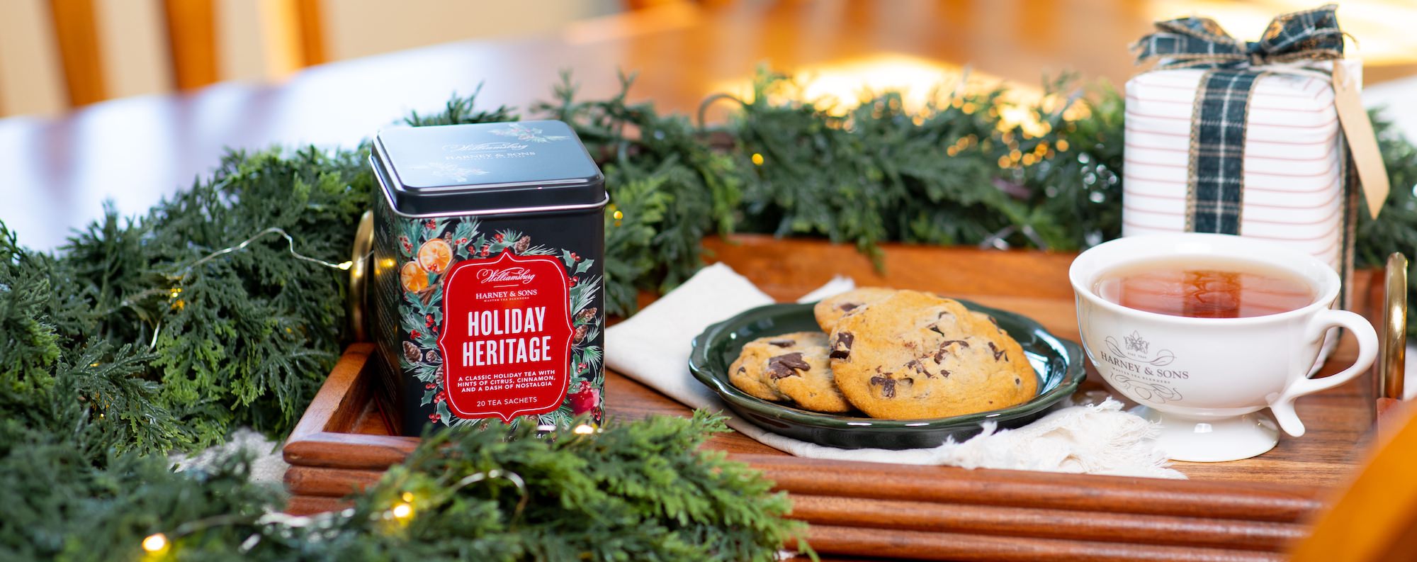 Tea tin with cookies and a cup of tea on a wooden tray, surrounded by greenery and holiday decorations.