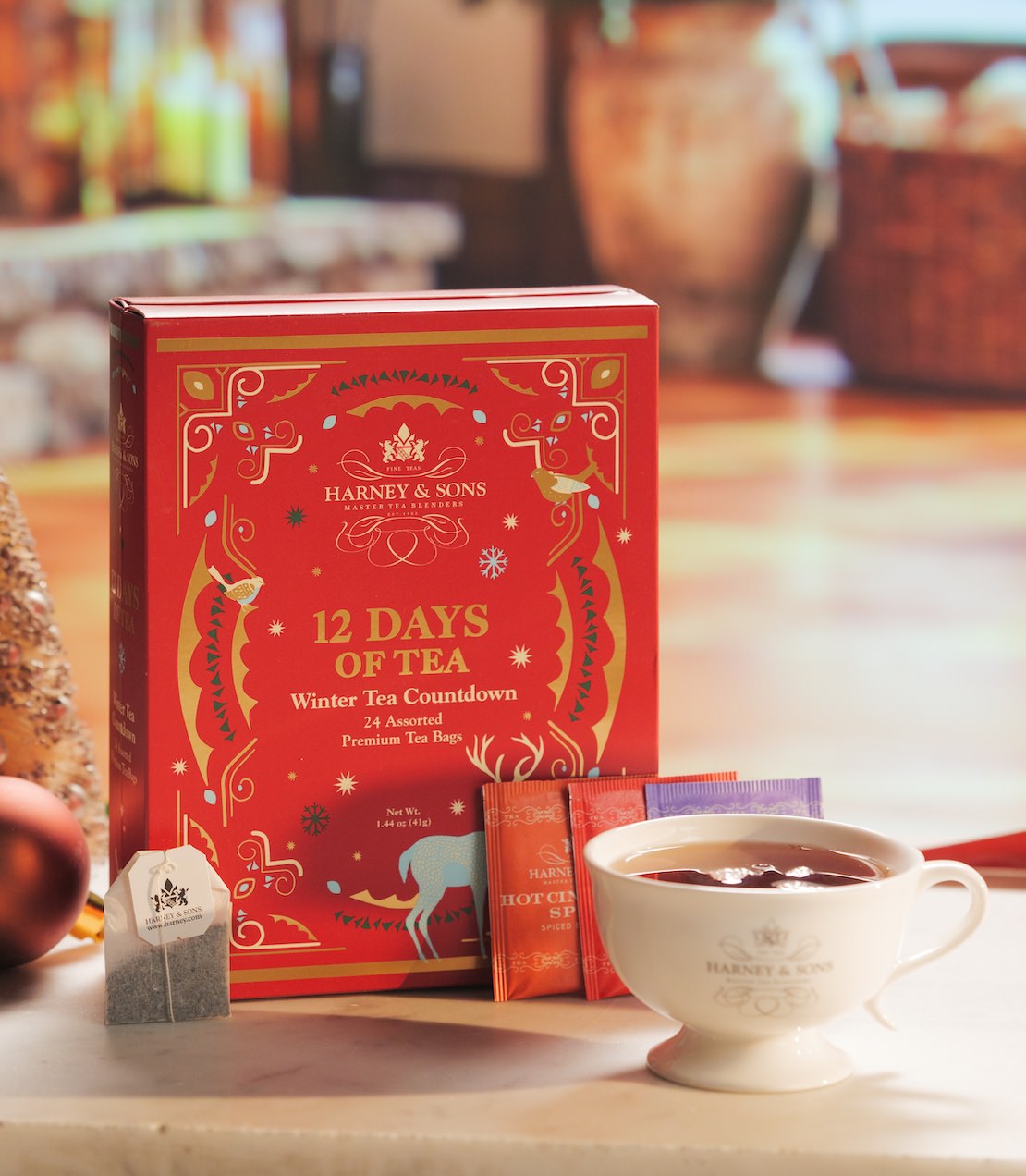 Harney & Sons '12 Days of Tea' box in red with gold decorative illustrations, shown on a tabletop next to a white teacup filled with tea and a tea bag, with a softly blurred warm indoor background.