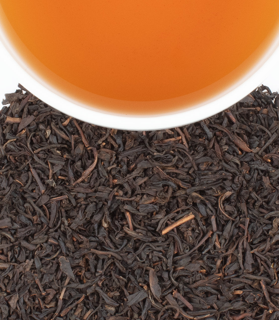 Close-up of loose dark tea leaves with a white cup filled with amber-orange tea at the top edge.