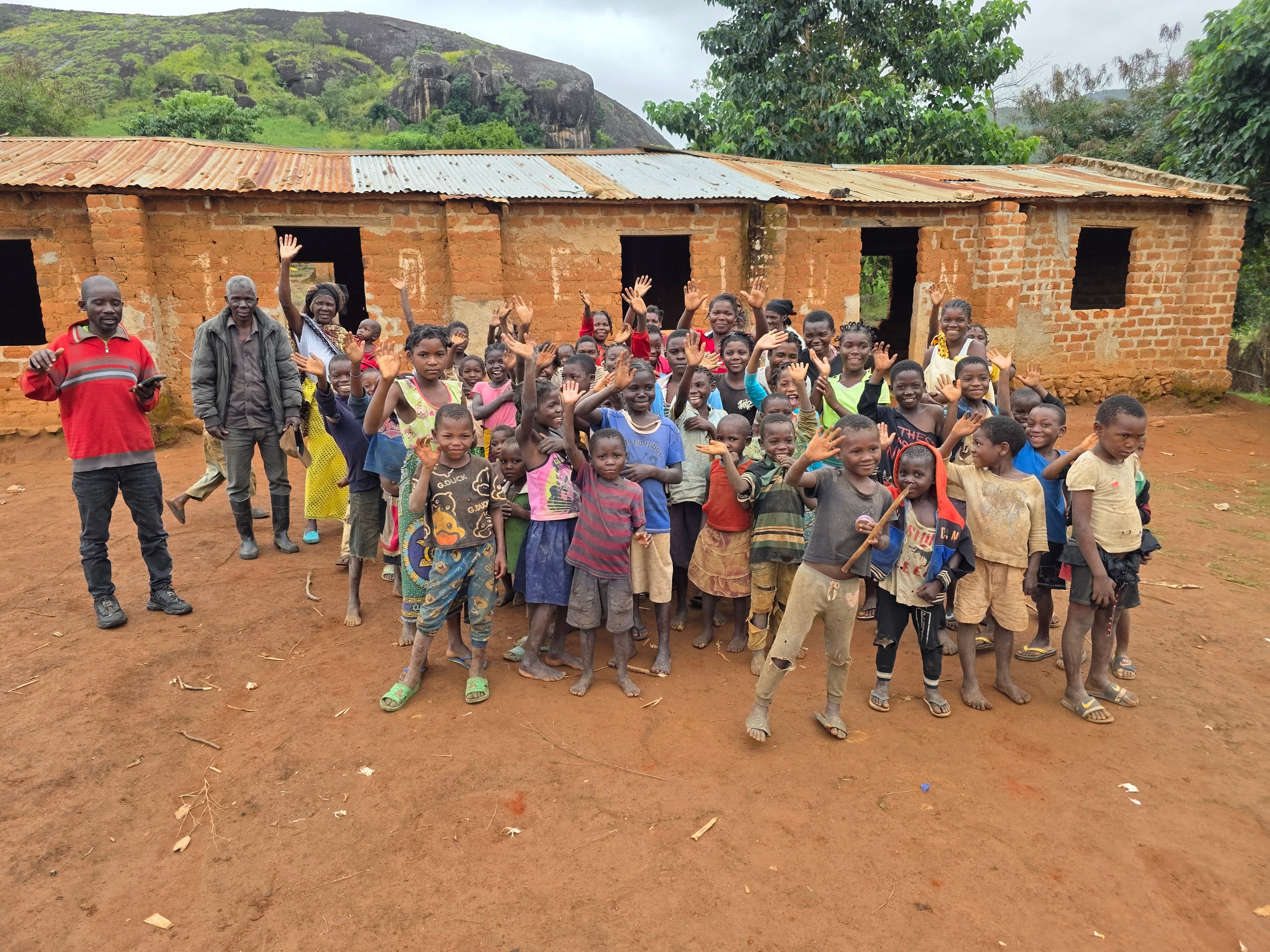 Children and adults gather outside a school in Mozambique, smiling and waving, showcasing community support for education.